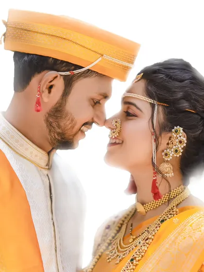 A close-up, candid shot of a Maharashtrian couple sharing a smile. The image highlights the intricate details of their wedding attire, from the bride's nath to the groom's mundavalya.