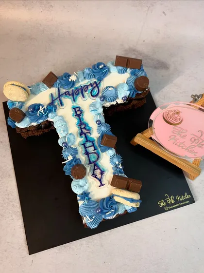 Here is an example of a letter cake, shaped into a 'T'. It's decorated with blue and white buttercream, chocolate chunks, and macarons, with 'Happy Birthday' piped down the center.