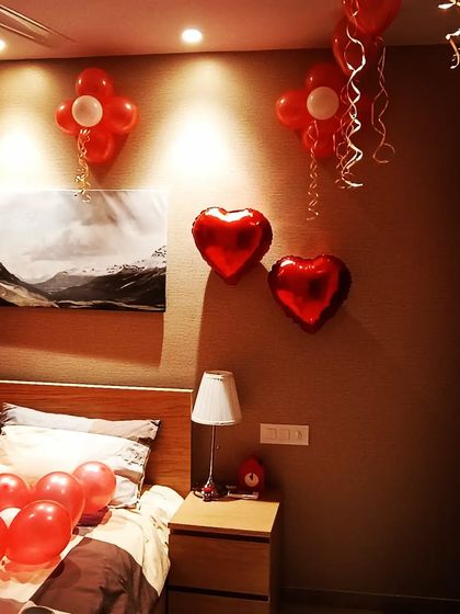 A full romantic room decoration surprise. The setup includes heart-shaped foil balloons, red balloons on the bed and ceiling, and soft lighting to create a cozy and loving atmosphere.