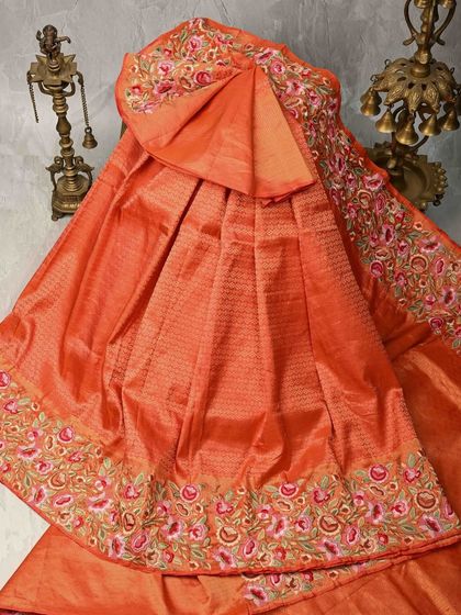 A vibrant orange silk saree with a rich, embroidered floral border. The bright color and intricate detailing make it a joyful and celebratory piece for any festive occasion.