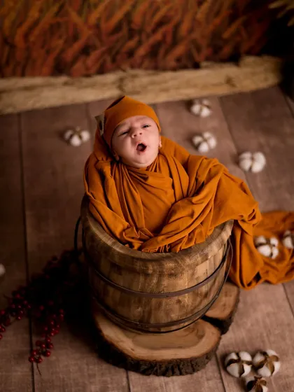 Our little pumpkin, perfectly ripe for cuddles. This autumn-themed shoot features a newborn wrapped in a warm orange swaddle, peeking out of a rustic wooden barrel surrounded by cotton props.