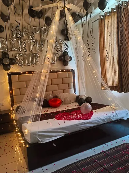 A surprise birthday room decoration with a canopy tent over the bed. The black and silver balloons, fairy lights, and rose petals create a stylish and romantic atmosphere.