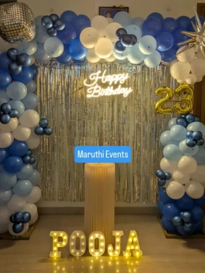A cool and stylish 23rd birthday celebration. The decor includes a blue, white, and silver balloon arch, a neon "Happy Birthday" sign, and light-up letters for the name.