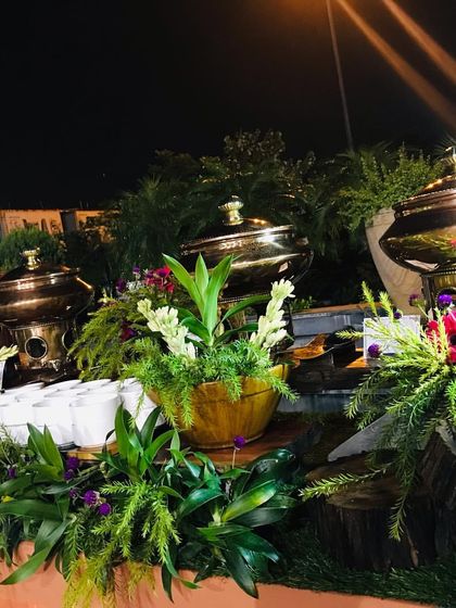 A close-up of an outdoor buffet with copper chafing dishes nestled in lush green and purple floral arrangements, set against a backdrop of a water feature.
