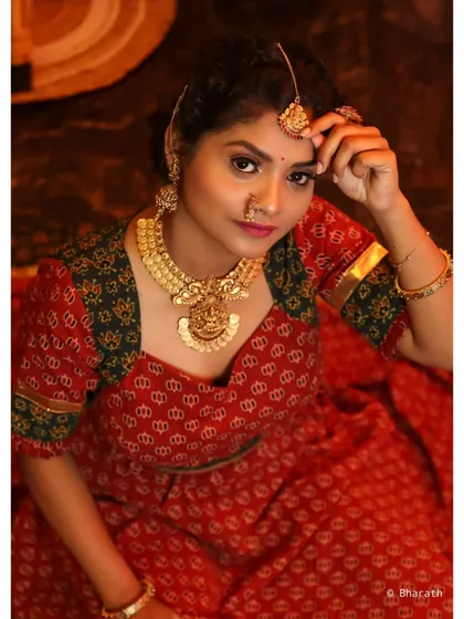 A direct and engaging portrait of a model in a traditional ensemble. She is adjusting her maang tikka, drawing the viewer's eye to the detailed jewelry and her confident gaze.