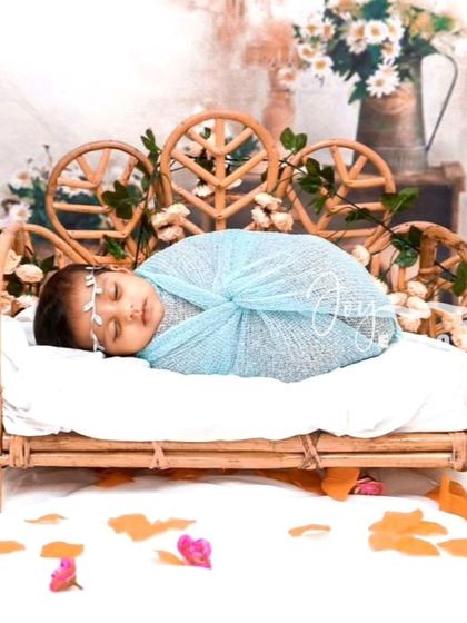 This serene newborn setup features a baby wrapped in light blue, resting on a miniature bamboo cot. The floral and leafy props create a peaceful, garden-like atmosphere for the portrait.