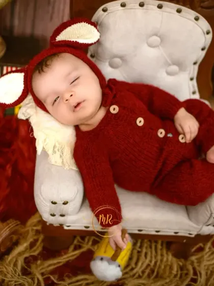 A sleeping newborn wearing a cute red outfit with mouse ears, resting on a miniature armchair.