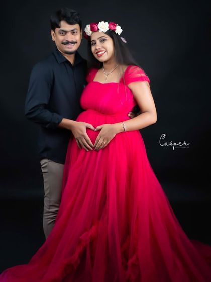 A sweet couple's portrait against a black background. The partner makes a heart shape with his hands on the baby bump, a classic and loving gesture.