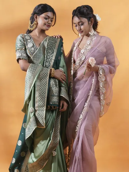 A lookbook image featuring two models styled in complementary designer sarees. One wears a sage green silk saree with intricate borders, while the other is in a delicate lavender organza saree with floral embroidery, highlighting my ability to style and shoot multiple looks together.
