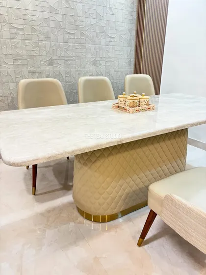 This modern dining set features a stunning Italian marble tabletop with softly rounded corners. The custom base is upholstered in a neutral quilted fabric with a brass-finished metal trim, perfectly complemented by elegant chairs with tapered wooden legs.