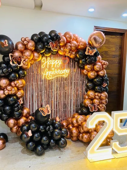 Celebrating a 25th anniversary with a stylish black and rose gold theme. This decor features a full ring of balloons, a shimmering fringe backdrop, a "Happy Anniversary" neon sign, and large light-up numbers to mark the milestone.