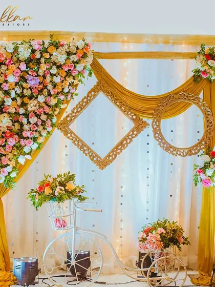 A creative and charming photo booth idea. The setup includes a decorative bicycle, floral arrangements, and ornate empty frames for guests to pose with.