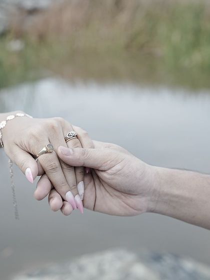 A close-up detail shot of a couple holding hands, showcasing their rings and connection, with the water in the background.