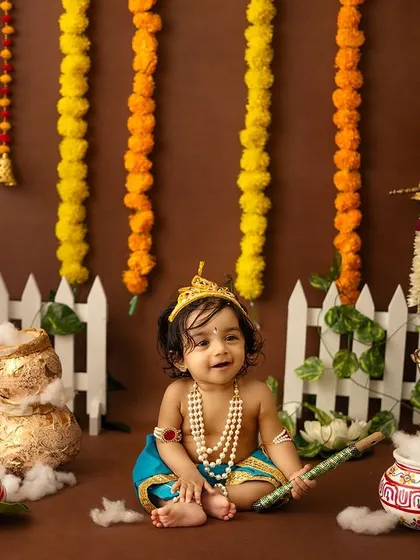 A happy baby Krishna smiles brightly in a setup decorated with marigold garlands and a white picket fence.
