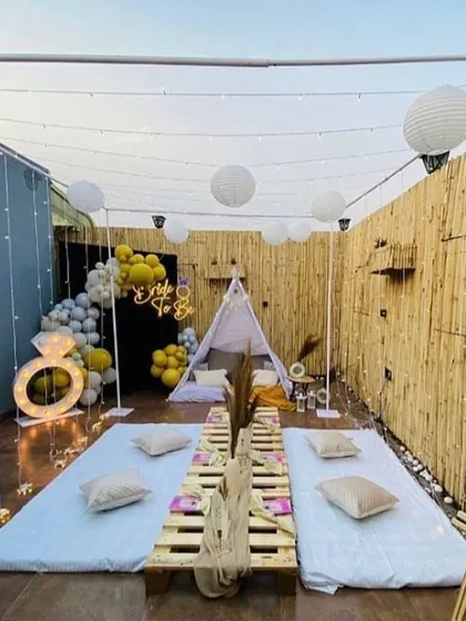 A full view of the bachelorette party setup on a terrace. The space is decorated with a 'Bride to Be' neon sign, a balloon arch, and overhead lighting with paper lanterns for a festive atmosphere.