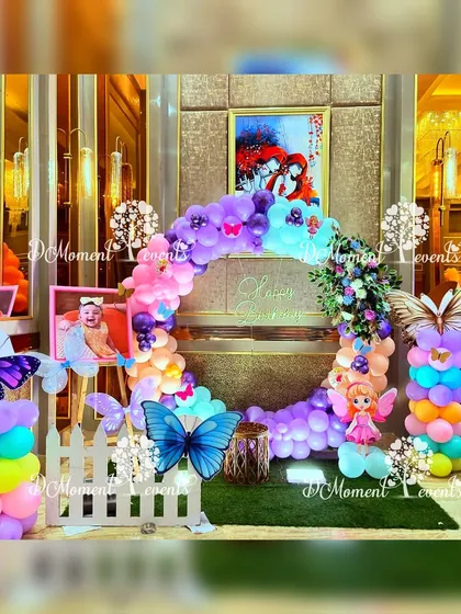 A circular balloon arch in vibrant rainbow colors, adorned with butterfly cutouts and a small white picket fence. This setup serves as a beautiful and cheerful photo spot.