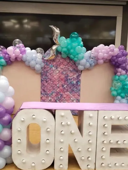 A first birthday setup with a mermaid-inspired color palette of purple, teal, and pink. The decor features large, light-up 'ONE' letters as the centerpiece.