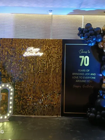 A sophisticated black and gold theme for a 70th birthday. This setup includes a gold sequin wall, a personalized sign, marquee numbers, and a stylish balloon garland.