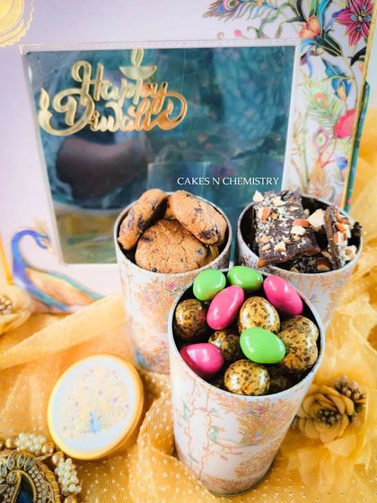 This hamper is designed with a beautiful peacock theme, containing jars of cookies, chocolate bark, and colorful coated almonds. It's a vibrant and delicious way to send your Diwali wishes.