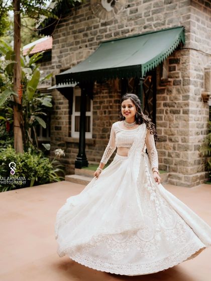 The bride twirling in her beautiful white lehenga at her engagement ceremony, captured in front of a rustic stone building.