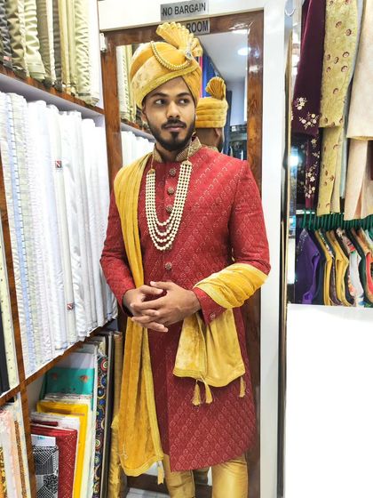 A groom looking dapper in a rich red textured sherwani. The outfit is completed with a golden turban and a matching stole, showcasing a classic and celebratory wedding style.