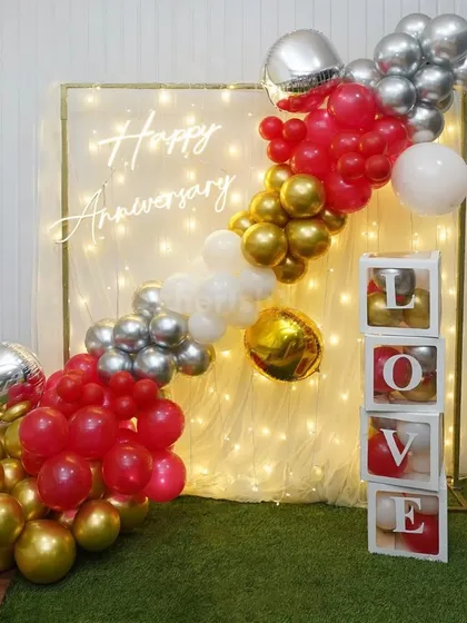 This is an example of an intimate anniversary decoration for a home celebration. It features a 'Happy Anniversary' neon sign on a fairy light backdrop, a vibrant balloon garland, and 'LOVE' display boxes for a personal touch.