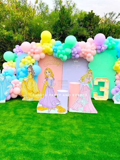 An outdoor Disney Princess themed birthday party. This beautiful setup on a green lawn features cutouts of various princesses against a pastel-colored backdrop and balloon garland.