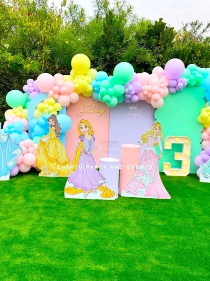 An outdoor Disney Princess themed birthday party. This beautiful setup on a green lawn features cutouts of various princesses against a pastel-colored backdrop and balloon garland.