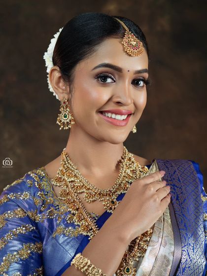 A close-up beauty shot of a South Indian bridal look. The focus is on the intricate gold jewelry and flawless makeup, with soft lighting that enhances the model's radiant smile.