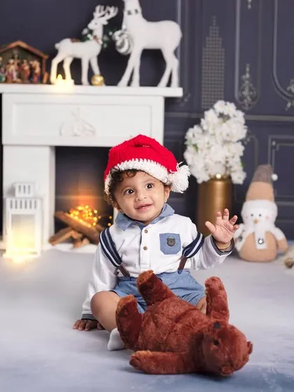 This little boy is ready for Christmas, playing with his teddy bear in front of a classic fireplace and decorated tree.
