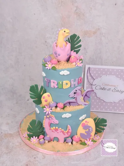 A two-tier dinosaur-themed cake for a little girl's 3rd birthday. The design features cute, friendly dinos in pastel colours, hatching from eggs and surrounded by tropical leaves.