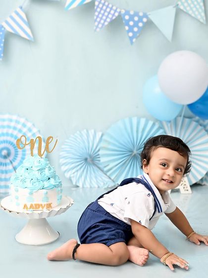 A classic blue and white cake smash for a baby boy's first birthday. The simple setup with paper fans and balloons creates a timeless look that lets his happy smile be the star.