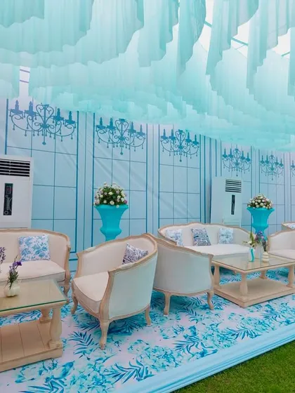 A luxurious lounge area at the Dior-themed Mehendi, featuring plush white sofas, custom-printed cushions, and a backdrop wall with elegant chandelier motifs.