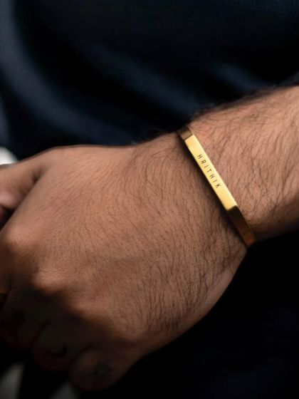 A close-up of a gold-plated cuff on a man's wrist, engraved with 'HRITHIK'. The warm tone of the gold stands out against the dark sleeve.