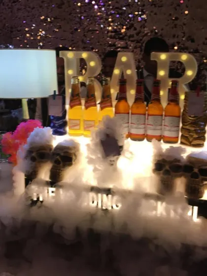 A spooky and fun theme for a party, this bar features skull-shaped glasses with drinks overflowing with smoke. The marquee 'BAR' lights and beer display complete this unique setup.