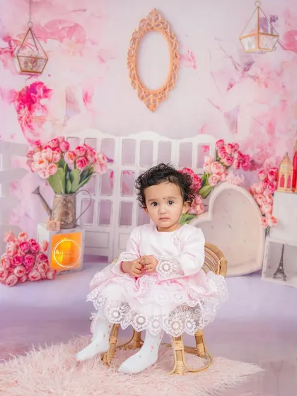 A sweet, seated portrait in a miniature wicker chair, set against a dreamy pink floral backdrop.