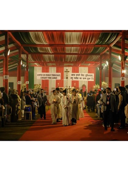 A Christmas mass procession walks down a red carpet inside a large tent, decorated in festive colors.