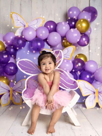 A sweet toddler dressed as a butterfly, smiling brightly for her milestone photoshoot. The purple and gold balloon arch and butterfly cutouts create a magical, whimsical setting.