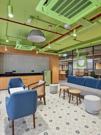 A breakout area with a fresh green ceiling and exposed red pipes. The space combines comfortable lounge seating with a small pantry, and uses patterned tiles to define the zone.