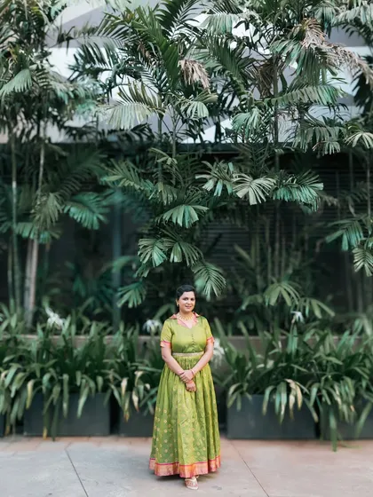A wide shot of a woman in a traditional green dress against a wall of lush greenery.
