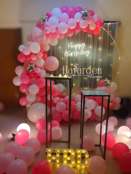 A romantic half-ring balloon setup in shades of pink. The fairy light curtain and scattered balloons on the floor create a dreamy atmosphere.