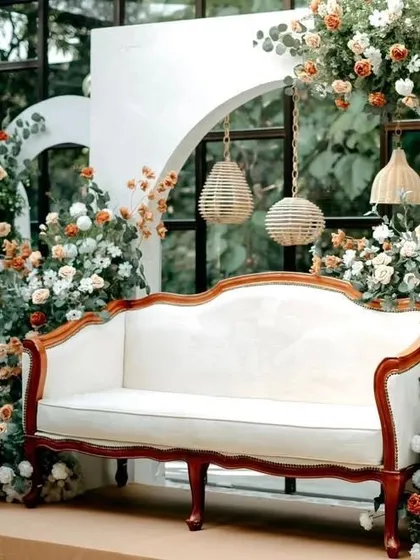 A modern photo backdrop with white arched panels and a vintage-style sofa. The setup is decorated with arrangements of white and rust-colored flowers, and rustic cane lamps hang within the arches.