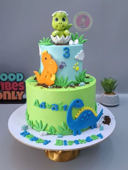 A two-tier dinosaur cake for Advait's 3rd birthday, with a baby dino hatching from an egg on top.