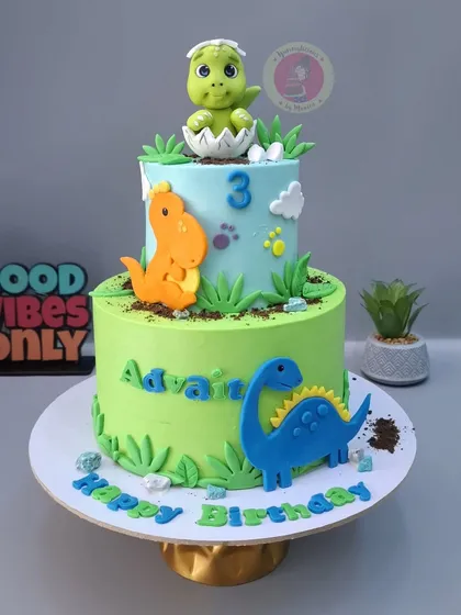 A two-tier dinosaur cake for Advait's 3rd birthday, with a baby dino hatching from an egg on top.