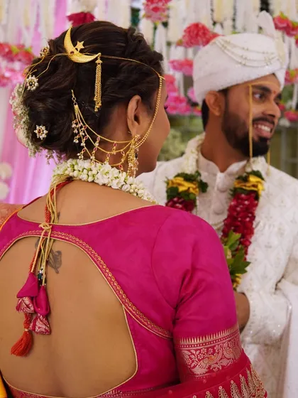A candid shot from behind, focusing on the intricate bridal hairstyle with a unique moon-shaped accessory and the beautiful back design of the blouse.