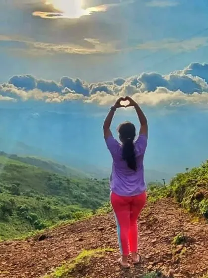 A trekker making a heart shape with her hands against the backdrop of the beautiful Chikmagalur hills.