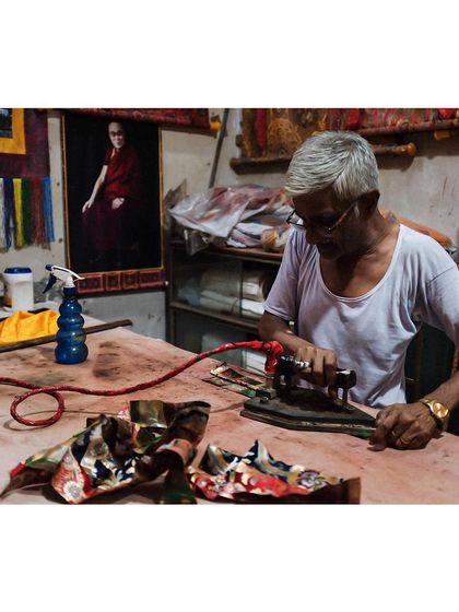 An elderly artisan with over two decades of experience, carefully ironing a colorful piece of cloth in his workshop. The portrait of the Dalai Lama in the background adds another layer to the story of his space.