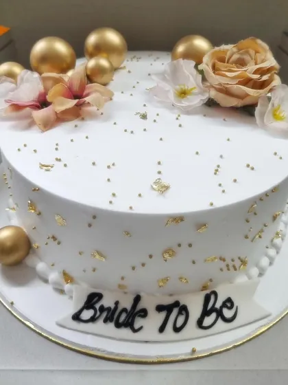 An elegant 'Bride to Be' cake in white, decorated with gold leaf, golden spheres, and delicate flowers. It's the perfect centerpiece for a bridal shower or bachelorette party.