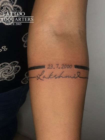 A minimalist armband tattoo that incorporates a special date, "23.7.2000," and the name "Lakshmi" in a fine-line script.