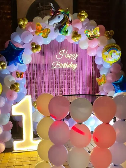 A unicorn-themed birthday setup with a balloon arch over a pink fringe curtain, a 'Happy Birthday' neon sign, and a light-up marquee number '1'.